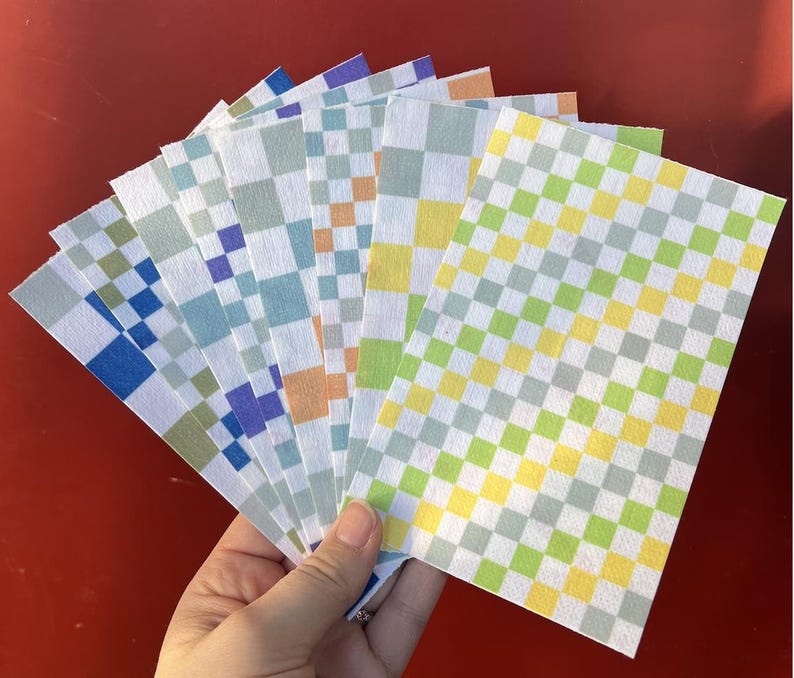 Puede incluir: Una mano sostiene un abanico de hojas de papel rectangulares con un patr&oacute;n de cuadros. Las hojas muestran varias combinaciones de colores, incluyendo azul, verde, amarillo y naranja, dispuestas en un dise&ntilde;o de cuadr&iacute;cula. El fondo es rojo liso.