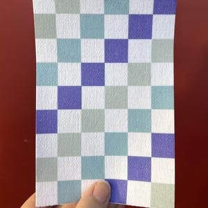 Puede incluir: Un trozo de papel rectangular con un patr&oacute;n de tablero de ajedrez. El patr&oacute;n consta de cuadrados en blanco, azul claro, verde salvia y morado. El papel tiene una superficie texturizada y lo sostiene una mano.