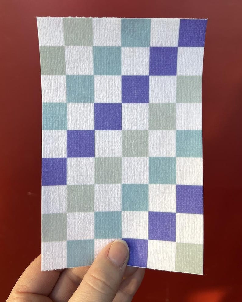 Puede incluir: Un trozo de papel rectangular con un patr&oacute;n de tablero de ajedrez. El patr&oacute;n consta de cuadrados en blanco, azul claro, verde salvia y morado. El papel tiene una superficie texturizada y lo sostiene una mano.