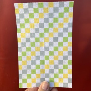 Puede incluir: Una hoja de papel rectangular con un patr&oacute;n de cuadros. El patr&oacute;n consiste en cuadrados alternos en amarillo, verde y gris. El papel se sostiene sobre un fondo rojo.