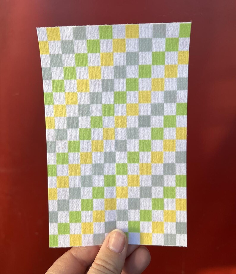 Puede incluir: Una hoja de papel rectangular con un patr&oacute;n de cuadros. El patr&oacute;n consiste en cuadrados alternos en amarillo, verde y gris. El papel se sostiene sobre un fondo rojo.