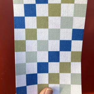 Puede incluir: Un trozo rectangular de papel con un patr&oacute;n de cuadros. El patr&oacute;n consiste en cuadrados azules, verdes y blancos. El papel est&aacute; sujeto por una mano sobre un fondo rojo.