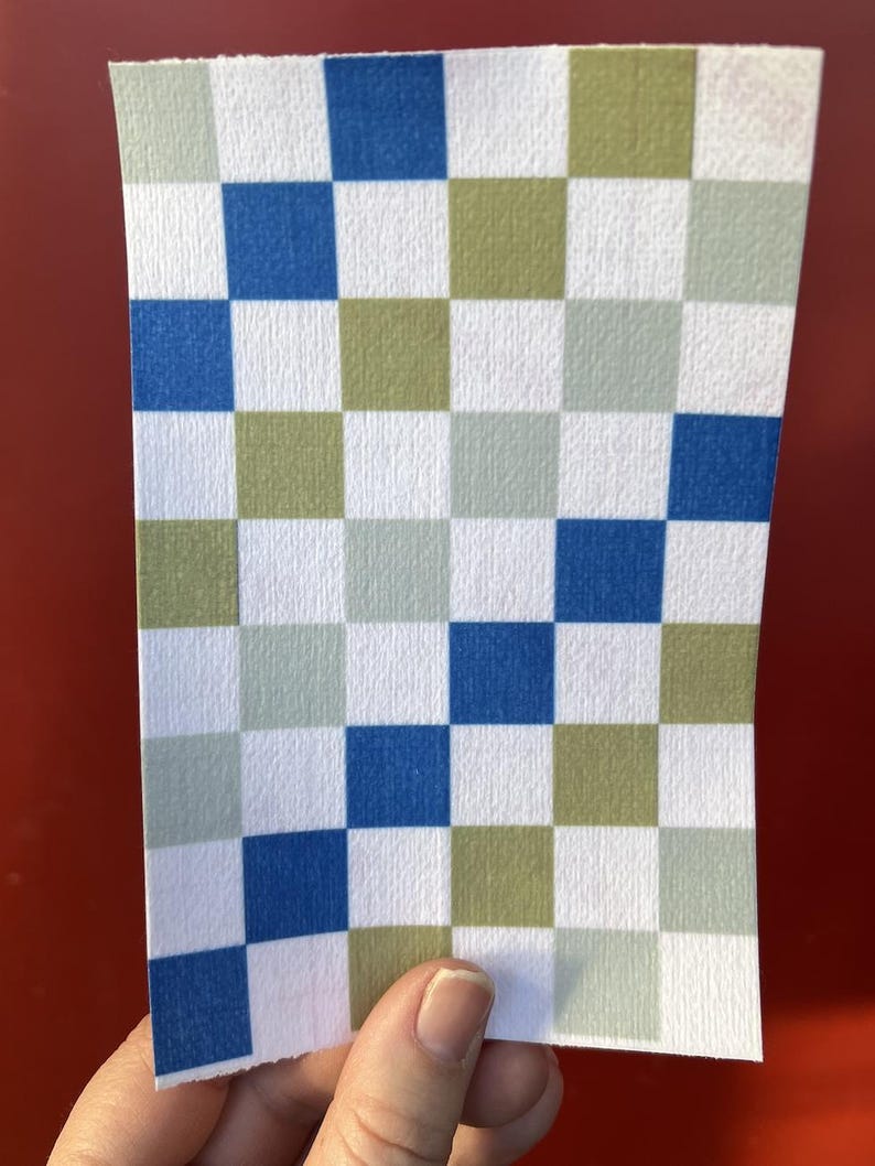 Puede incluir: Un trozo rectangular de papel con un patr&oacute;n de cuadros. El patr&oacute;n consiste en cuadrados azules, verdes y blancos. El papel est&aacute; sujeto por una mano sobre un fondo rojo.