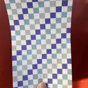 Puede incluir: Un trozo de papel rectangular con un patr&oacute;n de cuadros. El patr&oacute;n consiste en cuadrados alternos en blanco, azul, turquesa y gris. El papel est&aacute; sostenido por una mano sobre un fondo rojo.