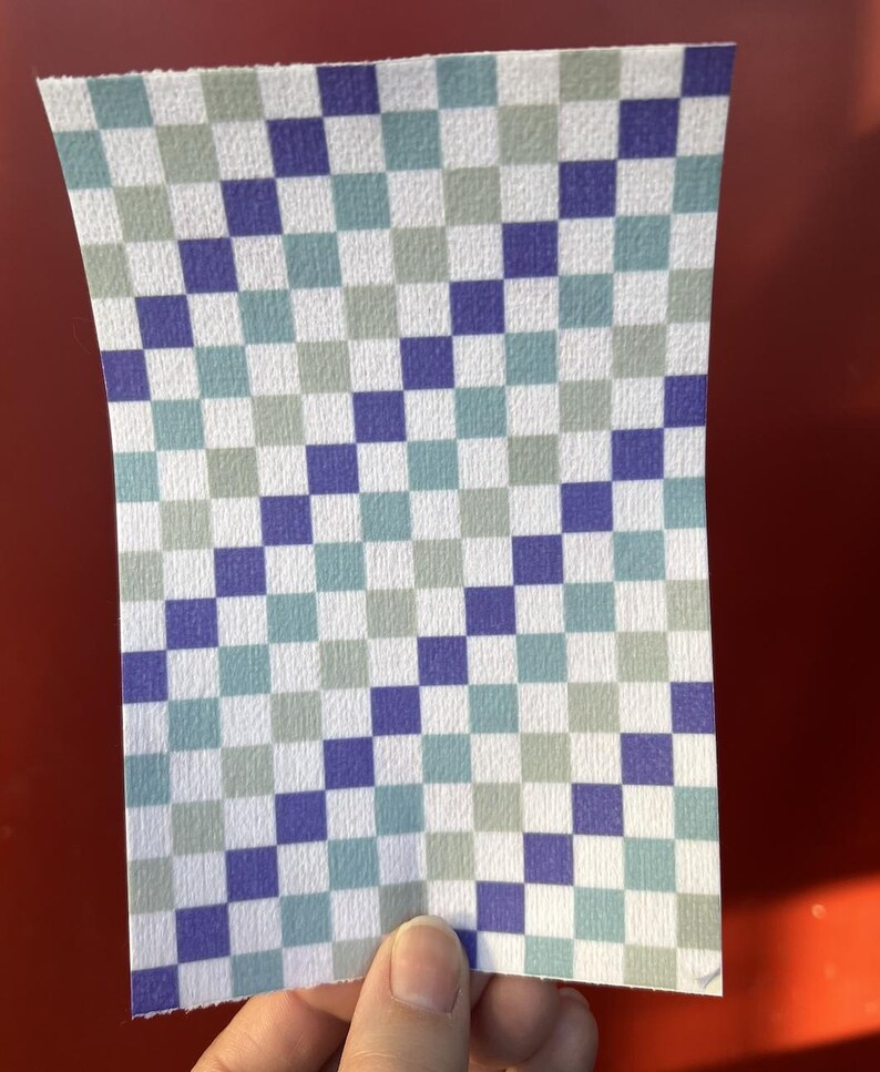 Puede incluir: Un trozo de papel rectangular con un patr&oacute;n de cuadros. El patr&oacute;n consiste en cuadrados alternos en blanco, azul, turquesa y gris. El papel est&aacute; sostenido por una mano sobre un fondo rojo.