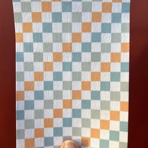 Puede incluir: Un trozo rectangular de papel con un patr&oacute;n de cuadros. El patr&oacute;n consiste en cuadrados alternos en blanco, azul claro, naranja y gris. El papel est&aacute; sujeto por una mano sobre un fondo rojo.