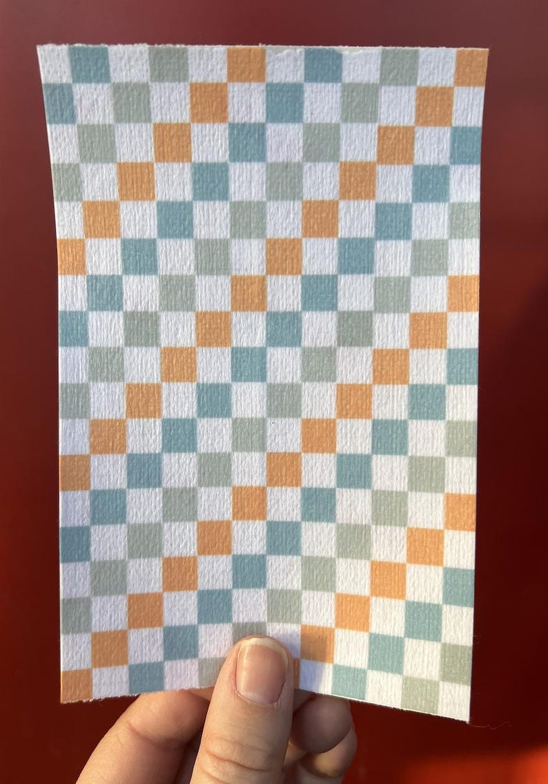 Puede incluir: Un trozo rectangular de papel con un patr&oacute;n de cuadros. El patr&oacute;n consiste en cuadrados alternos en blanco, azul claro, naranja y gris. El papel est&aacute; sujeto por una mano sobre un fondo rojo.