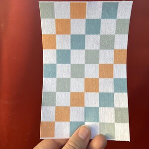 Puede incluir: Un trozo rectangular de papel con un patr&oacute;n de cuadros. El patr&oacute;n consiste en cuadrados alternos en blanco, naranja y tonos de azul y verde. El papel se sostiene sobre un fondo rojo.