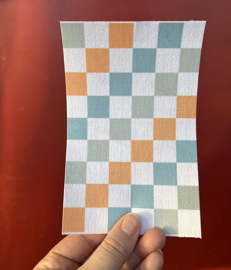 Puede incluir: Un trozo rectangular de papel con un patr&oacute;n de cuadros. El patr&oacute;n consiste en cuadrados alternos en blanco, naranja y tonos de azul y verde. El papel se sostiene sobre un fondo rojo.