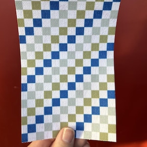 Puede incluir: Un trozo de papel rectangular con un patr&oacute;n de cuadros. El patr&oacute;n consta de cuadrados azules, verdes y blancos. El papel est&aacute; sujeto por una mano sobre un fondo rojo.