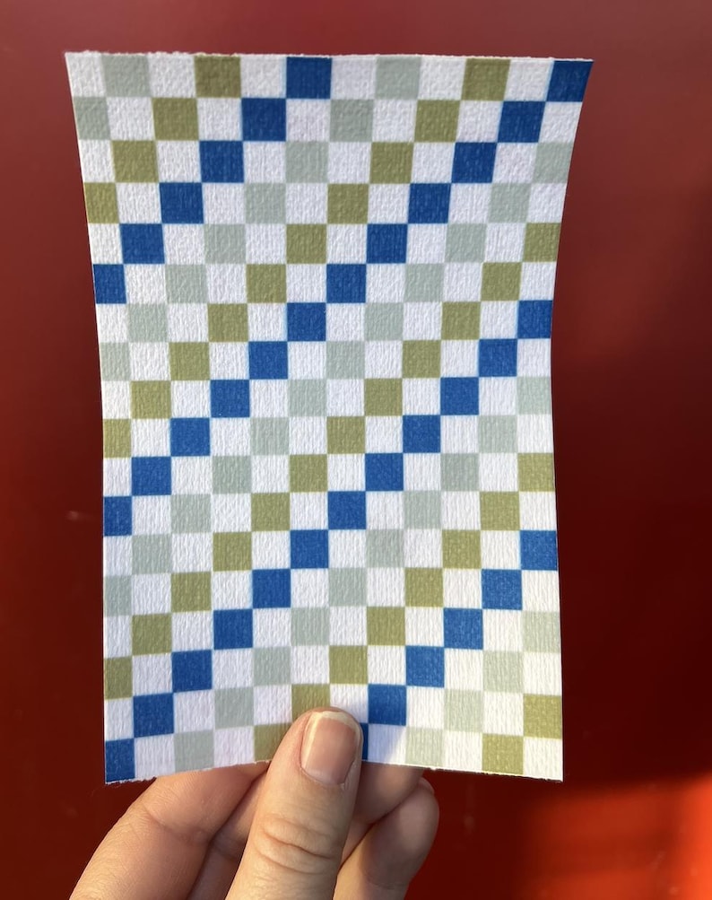 Puede incluir: Un trozo de papel rectangular con un patr&oacute;n de cuadros. El patr&oacute;n consta de cuadrados azules, verdes y blancos. El papel est&aacute; sujeto por una mano sobre un fondo rojo.