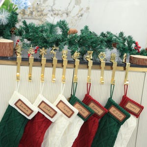 Può includere: Un set di calze natalizie verdi, rosse e bianche, ciascuna con un'etichetta con il nome. Le calze sono appese a un supporto in metallo dorato adornato con ornamenti festivi. Una ghirlanda di rami sempreverdi e bacche aggiunge un tocco festoso.