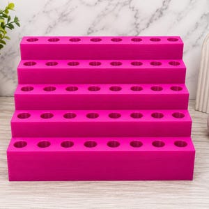 May include: A bright pink, tiered organizer with multiple circular holes. The organizer has four stepped levels, each with evenly spaced holes. The product is designed to hold small items, likely for crafting or beauty supplies.