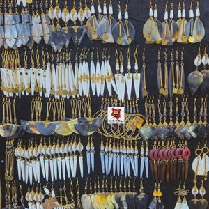 Puede incluir: Exhibición de pendientes artesanales variados. Los pendientes presentan diversas formas y materiales, con detalles blancos, grises y dorados. Muchos tienen ganchos dorados. Se exhiben sobre un fondo negro.
