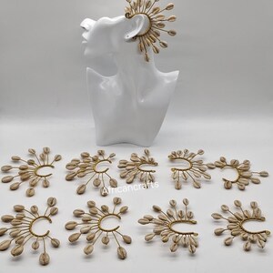 May include: A collection of ear cuffs featuring a gold-toned wire frame adorned with multiple white cowrie shells arranged in a fan-like pattern. One ear cuff is displayed on a white mannequin head, while nine others are arranged below it.