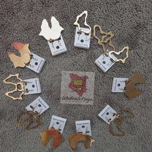 Puede incluir: Una colección de pendientes dorados en varias formas abstractas y geométricas, exhibidos en tarjetas blancas. Los pendientes están dispuestos sobre una superficie gris texturizada. El texto "Fashion Jewelry" es visible.