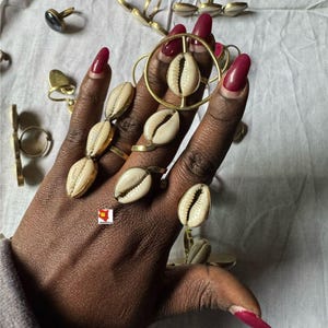 May include: A collection of gold-toned rings, some featuring cowrie shells. The rings are displayed on a hand with long, red nails. Other rings with various designs are scattered in the background, set against a white fabric.