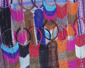 May include: A collection of beaded necklaces in various colors, including black, blue, red, orange, and pink. The necklaces have fringe detailing and are displayed on a purple and white patterned surface. Some necklaces have circular pendants.