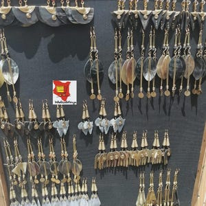 Puede incluir: Exhibición de varios pendientes hechos a mano. Los pendientes presentan una variedad de formas y materiales, incluyendo cuerno, metal y cuentas. Los diseños varían de simples a elaborados, con un enfoque en texturas naturales y tonos terrosos.