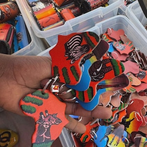 May include: A collection of keychains shaped like the African continent, featuring colorful animal illustrations. The keychains have red, blue, and green accents, with images of zebras, giraffes, and elephants. The keychains are attached to silver keyrings.