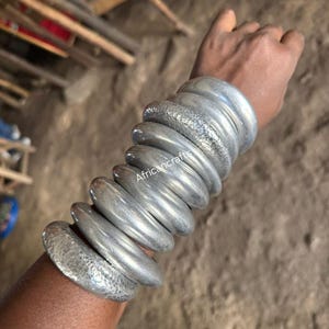Könnte beinhalten: Eine Ansammlung silberfarbener Armreifen, einige mit strukturierter Oberfläche, sind auf einem Arm gestapelt. Die Armreifen variieren in Breite und Design und erzeugen einen geschichteten Effekt. Das Bild ist in natürlichem Licht aufgenommen.