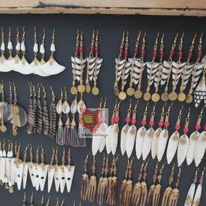 May include: A collection of handcrafted earrings with various designs. Some earrings feature white, fan-shaped pendants, while others have brown, striped, or shell-like elements. Many have gold-colored metal accents and red or brown beads. The earrings are displayed on a black surface.