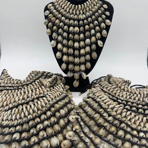 May include: A display of handcrafted shell necklaces and matching accessories. The necklaces are made of small, cream-colored shells strung together with black cord. One necklace is displayed on a black mannequin head, while others lay flat.