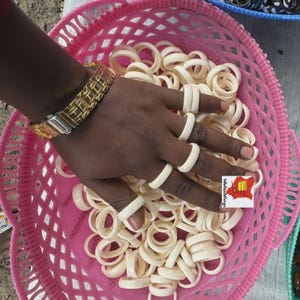 Op de afbeelding: Een hand met een gouden horloge en meerdere witte ringen rust in een roze mand gevuld met meer witte ringen. De ringen hebben een eenvoudig, afgerond ontwerp. De mand is gemaakt van geweven plastic.