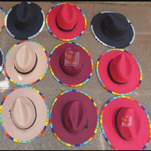 May include: A selection of fedora hats in colours such as black, red, and beige. Each hat has a colourful beaded trim around the brim. Some hats have a logo on the crown.