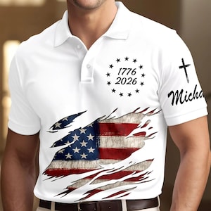 May include: White polo shirt featuring a distressed American flag design. The shirt has a torn effect revealing the flag's stars and stripes. The text "1776 2026" is above the flag, and a cross with the name "Micha" is on the sleeve.