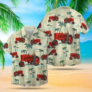 Puede incluir: Una camisa con botones de manga corta con un patrón repetido de tractores rojos y palmeras sobre un fondo beige claro. La camisa está en una playa con palmeras y un cielo azul.