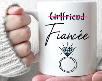 May include: White ceramic mug with the word "Fiancée" in a script font, with a diamond ring graphic. The word "Girlfriend" is crossed out above "Fiancée". The mug is held by a person wearing a white sweater.