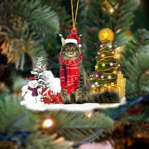May include: A Christmas ornament featuring a tabby cat wearing a Santa hat and a red scarf. The cat is surrounded by a small Christmas tree, a snowman, and wrapped gifts. The ornament hangs from a gold string.