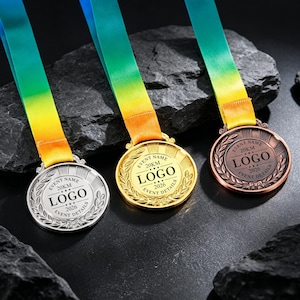 Könnte beinhalten: Drei kreisförmige Metallmedaillen in Silber, Gold und Bronze mit einem Lorbeerkranzrand und dem Text EVENT NAME, 20KM, LOGO, 2026, EVENT DETAILS. Jede Medaille ist an einem Farbverlaufsband befestigt, das von Blau zu Gelb übergeht.