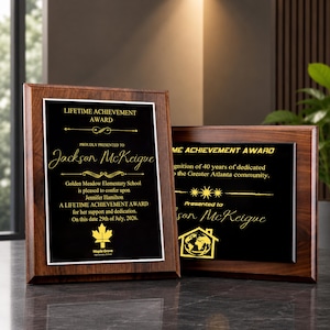 Puede incluir: Dos placas rectangulares de madera con marcos de color marrón oscuro y placas centrales negras con texto dorado. La placa de la izquierda dice Lifetime Achievement Award, Jackson McKeigue, Golden Meadow Elementary School y 29 de julio de 2026.