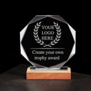 Puede incluir: Trofeo de cristal octogonal con centro negro, con el texto "YOUR LOGO HERE" dentro de una corona de laurel y "Create your own trophy award". El trofeo se apoya sobre una base de madera.