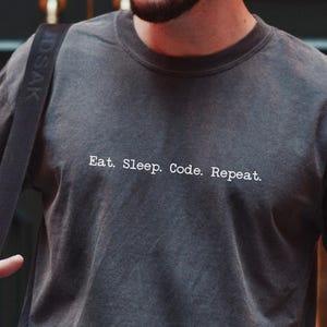 Op de afbeelding: Donkergrijs t-shirt met de witte tekst "Eat. Sleep. Code. Repeat.". Het shirt heeft een licht vervaagde uitstraling. Een zwarte schoudertas is zichtbaar. De persoon draagt een horloge en een armband.