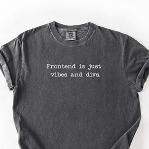 Op de afbeelding: Een donkergrijs t-shirt met de witte tekst "Frontend is just vibes and divs". Het shirt heeft een ronde hals en opgerolde mouwen. De stof heeft een licht vervaagde, vintage uitstraling.