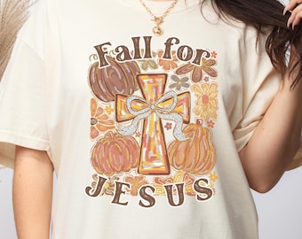 May include: Ivory t-shirt with a fall-themed graphic. The design features pumpkins, flowers, and a cross with a bow, along with the text "Fall for Jesus". The shirt is a casual, short-sleeved style.