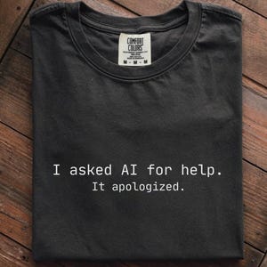 Könnte beinhalten: Schwarzes T-Shirt mit dem weißen Text "I asked AI for help. It apologized.". Das T-Shirt ist auf einer Holzoberfläche gefaltet. Das Etikett der Marke Comfort Colors ist am Halsausschnitt sichtbar.