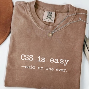 Op de afbeelding: Espresso-kleurig T-shirt met de tekst "CSS is easy - said no one ever." Het shirt is van het merk Comfort Colors, maat S-P-CH. Een zilveren ketting met een kleine hanger ligt over het shirt. Een bruine hoed is gedeeltelijk zichtbaar.