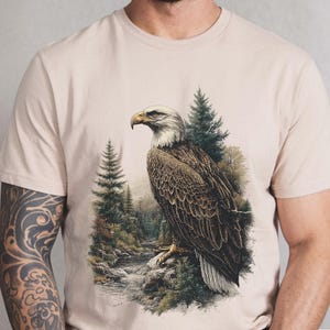 May include: Ivory-colored t-shirt featuring a detailed illustration of a bald eagle perched on a rock. The eagle is set against a backdrop of a forest and a flowing stream. The shirt has a classic crew neck and short sleeves.