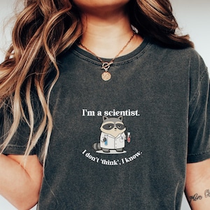 Peut inclure: T-shirt gris foncé avec un raton laveur de dessin animé portant une blouse de laboratoire et tenant des tubes à essai. Le texte dit : « Je suis un scientifique. Je ne pense pas, je sais. » Le t-shirt est de style décontracté.