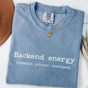 May include: A blue jean Comfort Colors t-shirt with the text "Backend energy: invisible, critical, underpaid." A gold necklace with a pendant is draped across the shirt. The shirt is folded on a white surface.