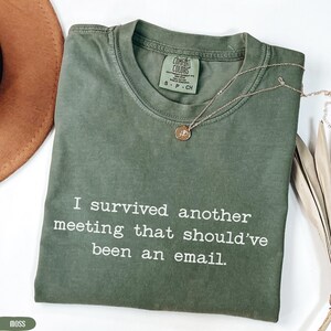 Puede incluir: Una camiseta verde musgo con el texto "I survived another meeting that should've been an email". Un collar dorado con un colgante descansa sobre la camiseta. La camiseta está etiquetada como "Comfort Colors" y es talla S-P-CH.
