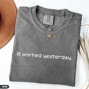 Puede incluir: Una camiseta gris Comfort Colors con el texto "It worked yesterday." Un collar plateado con un colgante circular descansa sobre la camiseta. Un sombrero marrón y hojas decorativas son visibles en el fondo.