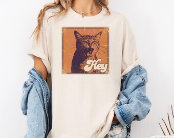 Puede incluir: Camiseta color marfil con un gráfico de estilo vintage de un gato con la boca abierta, dentro de un cuadrado marrón. La palabra "Hey" está escrita debajo del gato. La camiseta tiene mangas enrolladas.