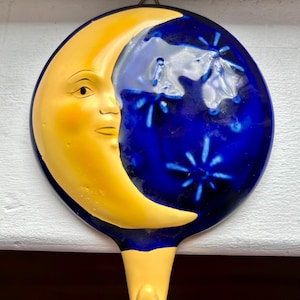 Puede incluir: Un colgador de pared de cerámica con una luna creciente con una cara amarilla y un fondo azul oscuro con estrellas blancas. El gancho es amarillo y está diseñado para colgar en una pared.