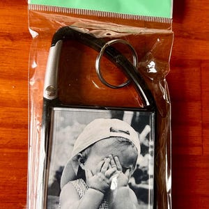Könnte beinhalten: Ein Acryl-Foto-Schlüsselanhänger mit Karabinerhaken. Der Schlüsselanhänger enthält ein Schwarz-Weiß-Foto eines Kindes mit Mütze. Die Verpackung trägt die Aufschrift "acrylic photo keychain" und misst 5 cm x 7,3 cm.