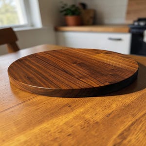 Puede incluir: Una tabla de cortar redonda de madera con un acabado marrón oscuro intenso. La tabla tiene una superficie lisa y un patrón de vetas de madera natural. Los bordes son ligeramente redondeados. La tabla tiene unos 30 cm de diámetro.